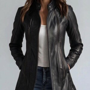 Puede incluir: Chaqueta de cuero negra con cuello alto y corte entallado. La chaqueta presenta cierre de cremallera, múltiples bolsillos y un diseño largo y elegante. La modelo lleva un top blanco y vaqueros azules.