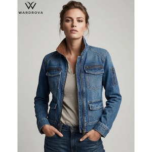 May include: A blue denim jacket featuring a stand-up collar with a brown inner lining, a full-length front zip, two chest pockets with zips, two lower flap pockets, and a small zipped utility pocket on the left sleeve, worn over a striped shirt.