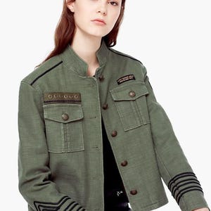 May include: An olive green military-style jacket featuring a mandarin collar, front button closure, and two chest pockets. The jacket includes black braided trim on the cuffs and shoulder, with a small rectangular patch reading ARMY CASUAL WEAR BARCELONA 1984.