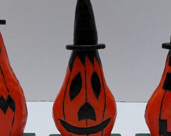 May include: Three orange pumpkin-shaped figures with black carved facial features and black pointed hats. Each figure is mounted on a square dark green base. The pumpkins display different expressions, including jagged, smiling, and blocky mouths.