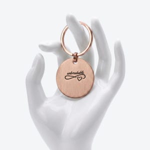 May include: A rose gold circular keychain featuring a brushed metal finish. The front of the disc is engraved with the word unbreakable in a cursive script, positioned directly above an infinity symbol that incorporates a small heart shape.