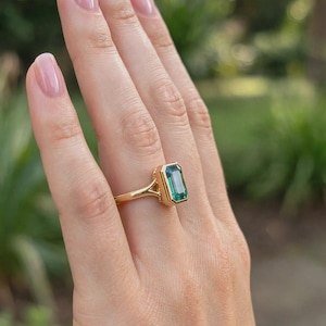Könnte beinhalten: Ein Goldring mit einem rechteckigen, smaragdgeschliffenen grünen Edelstein in einer Zargenfassung. Die Ringschiene ist schmal und glatt und verbindet sich mit der Fassung durch ein geteiltes Design. Der Edelstein zeigt eine leuchtend grüne Farbe.