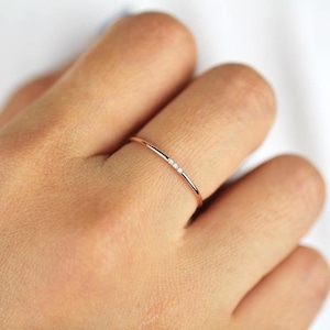 Pode incluir: Um delicado anel de ouro rosa com três pequenas pedras preciosas transparentes. O anel tem um design minimalista com uma banda fina. O anel é mostrado num dedo. Adequado para uso diário.