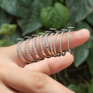 Puede incluir: Una mano con varios anillos de plata con piedras preciosas transparentes. Los anillos varían en ancho, con tamaños etiquetados de 1,00 mm a 3,00 mm. El fondo es verde borroso.