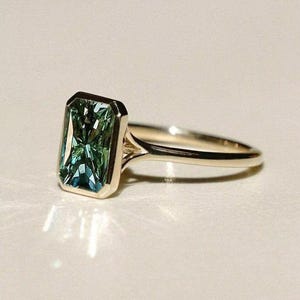Könnte beinhalten: Ein goldener Ring mit einem rechteckigen, smaragdgeschliffenen Edelstein in Grün- und Blautönen. Der Edelstein ist in einer goldenen Lünettenfassung gefasst, und das Band ist aus poliertem Gold. Der Ring wird auf einer hellen Oberfläche gezeigt.