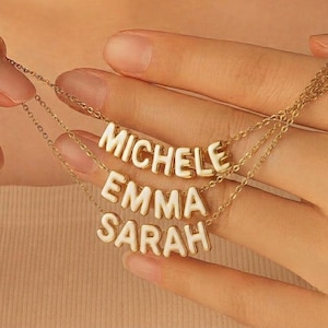 May include: Three gold-coloured necklaces, each bearing a name in white block letters: "MICHELE", "EMMA", and "SARAH". The necklaces are held in a hand, highlighting the fine chain and pendant design. The background is a soft, neutral tone.