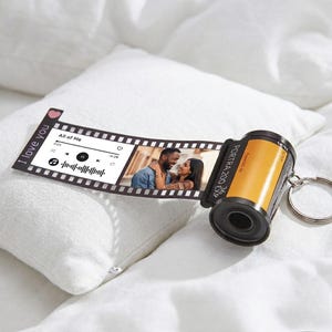 May include: A camera film roll keychain featuring an extended strip of film. The strip displays a music player interface with the text All of Me and a photograph of a couple. The film canister is yellow and black with the text PORTRA 200 36 EXP.