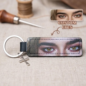 May include: A rectangular leather keychain featuring a printed close-up of human eyes with hazel irises. The keychain has a silver metal split ring attached to a small silver cross charm. Text above the keychain reads CUSTOM EYES.