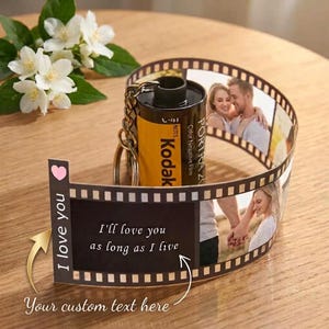 Puede incluir: Un llavero de cartucho de película Kodak Portra 200 unido a una tira retráctil de película fotográfica. La tira muestra fotografías de una pareja y el texto I love you as long as I live, junto a un icono de un corazón rosa.