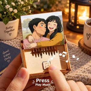 Puede incluir: Una caja de música de madera con una foto cuadrada de una pareja abrazándose en la parte superior. El panel frontal muestra un diseño de onda sonora recortado y un pequeño botón cuadrado con un icono de nota musical. El texto dice Press Play Music.