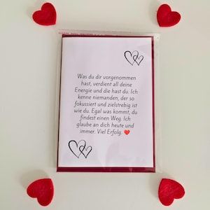 May include: A white greeting card with black text in German, featuring two black heart illustrations and a small red heart icon. The card is enclosed in a clear plastic sleeve and is surrounded by four small red felt hearts at the corners.