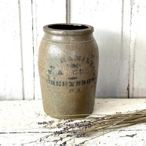 May include: A stoneware crock with a cylindrical shape and a dark interior. The crock has a light brown color with black lettering that reads "Greensboro". Dried lavender sprigs are in the foreground.