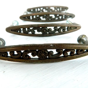 May include: Three elongated oval drawer pulls made of aged bronze-toned metal. Each handle features an openwork design with scrolling filigree patterns and a central circular floral motif. The pulls are arranged in a row, showing mounting hardware.