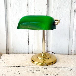 May include: A banker's lamp featuring a polished gold-tone metal base and a cylindrical stem. The lamp has a vibrant green glass shade with a curved profile and a black pull chain switch hanging from the underside for manual operation.