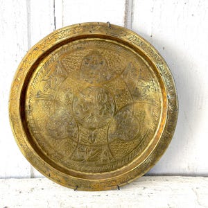 May include: A round, antique brass tray with intricate etched designs. The tray features a detailed central pattern surrounded by a decorative border. The aged brass has a warm, golden hue, suggesting its vintage origin. A decorative piece for serving or display.