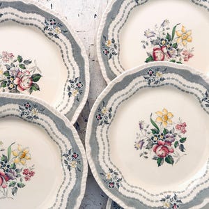 May include: A set of three cream-colored ceramic plates with scalloped edges. Each plate features a wide, textured gray border with a delicate floral pattern and a central bouquet of colorful flowers including roses and daffodils.