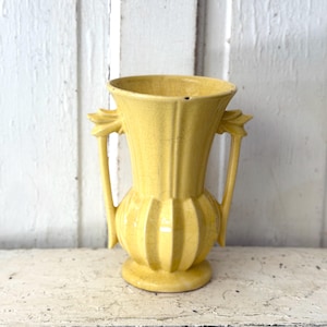 May include: A yellow ceramic vase with a flared rim, a bulbous base, and two vertical handles on the sides. The surface features a distinct crackle glaze finish throughout, with vertical ridges running along the body and neck of the vessel.