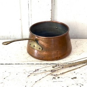 May include: A round copper saucepan with a metallic interior and a long, flat brass handle attached to the side with three rivets. The exterior surface of the pot shows a warm, polished copper finish with subtle signs of age and use.
