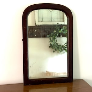 May include: An arched wall mirror with a dark brown wooden frame. The reflective glass surface shows a partial reflection of a white wall, a dark wooden board with white knobs, and a green leafy wreath hanging in the background.