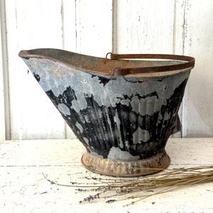 May include: A vintage metal coal scuttle with a handle. The scuttle has a weathered, distressed appearance with gray and black paint. The base is a lighter color, and the handle is a rusty brown. The scuttle is on a white surface.