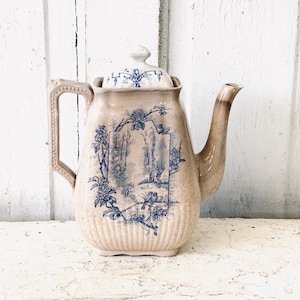 May include: A ceramic teapot with a beige speckled finish and a blue transferware print featuring a landscape with trees and birds. The teapot has a square-shaped body with vertical ribbing at the base, a curved handle, a spout, and a matching lid.