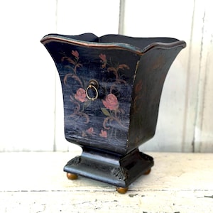 May include: A black, vintage-style wooden planter with a flared top and a square base. The planter features hand-painted pink roses and gold accents. A small, gold ring pull is on the front. The planter sits on four small, gold-colored feet.