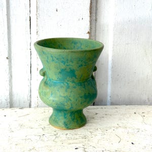 May include: A green ceramic vase with a speckled turquoise and green design. The vase has a flared top, a bulbous midsection with small handles, and a pedestal base. The background is a white, weathered wooden surface.