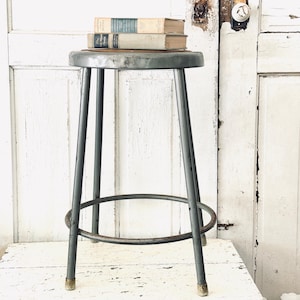 May include: A vintage gray metal stool with a round seat and four straight legs connected by a circular footrest. Two hardcover books are stacked on the seat. The stool has a weathered finish with visible signs of wear and metallic texture.