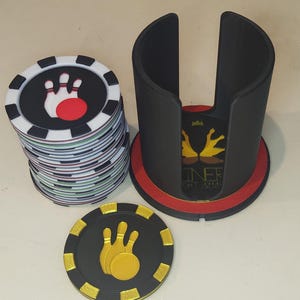Puede incluir: Fichas de póquer con temática de bolos con un diseño negro, blanco, rojo y verde. Un soporte para fichas negro con base roja y detalles dorados está junto a las fichas. También se ve una sola ficha con un diseño dorado de bolos.
