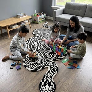 Op de afbeelding: Een groot vloerkleed in de vorm van een slang, voorzien van een contrastrijk zwart-wit patroon met dambord- en wervelende ontwerpen. Het kleed ligt op een houten vloer en biedt een ondergrond om te spelen met diverse kleurrijke speeltjes.