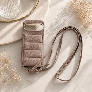 Puede incluir: Una funda para teléfono de color topo con un diseño acolchado texturizado y una correa a juego. La funda tiene forma rectangular con un recorte para la cámara y una correa que se puede llevar alrededor del cuello o del hombro. La funda está hecha de un material suave similar al cuero.