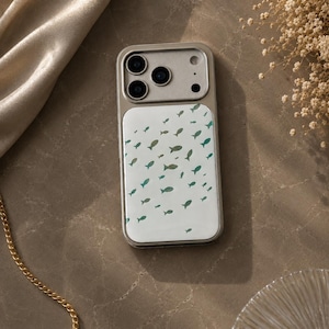 May include: A silver phone case with a white backing featuring a pattern of small, teal-colored fish. The case has a metallic frame and a camera module with three lenses. The phone case is on a marble surface.