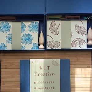 Pode incluir: Um conjunto de caixas azuis contendo kits de encadernação japonesa. Os kits incluem papel estampado, ferramentas e um furador de madeira. A caixa em primeiro plano tem o texto "KIT Creativo Rilegatura Giapponese".
