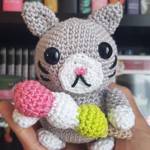 May include: A gray crochet cat figurine with black eyes, whiskers, and a pink nose. The cat holds a colorful ice cream cone with pink, white, and green sections. The handmade cat is a whimsical decorative item.