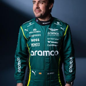 Może przedstawiać: Zielono-czarny kombinezon wyścigowy z żółtymi akcentami. Kombinezon posiada logotypy sponsorów, w tym "aramco", "MAADEN" i "cognizant". Na kombinezonie wydrukowano nazwisko "ALONSO". Na nadgarstku widoczny jest zegarek.
