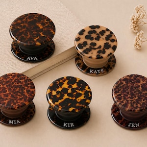 May include: Five personalized phone grips with a tortoiseshell pattern. Each grip has a different name printed on the base: AVA, KATE, MIA, KIR, and JEN. The grips are black and brown with a glossy finish.