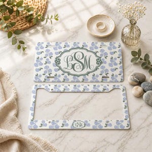 May include: A license plate and frame set featuring a floral hydrangea pattern in light blue and white. The license plate has a central monogram design in a darker blue-green color. The frame also has the floral pattern and a matching monogram.