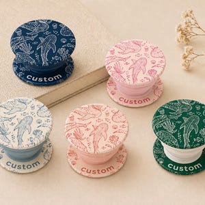 May include: A collection of custom phone grips with marine life designs. The grips feature various colors, including navy blue, pink, and green, with white illustrations of sharks, fish, and coral. Each grip has the word "custom" printed on it.