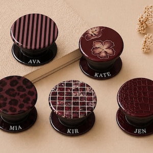 May include: A collection of personalized phone grips in various designs. The grips feature patterns like stripes, leopard print, floral, mosaic, and a crocodile texture. Each grip has a name printed on the base in white. The colors are shades of burgundy and black.