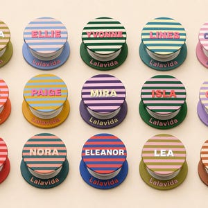 May include: A collection of fifteen personalized phone grips with horizontal striped designs in various colors. Each grip features a different name in bold letters, with the brand name "Lalavida" printed on the base.