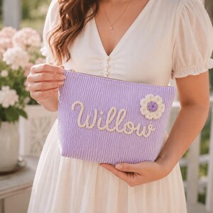 Könnte beinhalten: Eine hellviolette, plissierte Reißverschlusstasche mit dem Namen Willow in weißer Schreibschrift und einer kleinen, weiß-violetten Häkelblumenapplikation in der oberen rechten Ecke der Stoffoberfläche.