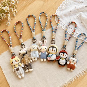 Puede incluir: Siete llaveros de peluche con forma de gato, cebra, perro, conejo y dos pingüinos, cada uno unido a un lazo de tela trenzada de colores con un envoltorio de cordón color canela. Las figuras visten diversos atuendos, incluyendo petos y vestidos.