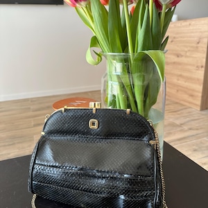 Może przedstawiać: Czarna, teksturowana torebka ze złotym łańcuszkiem leży na ciemnej powierzchni. Torebka ma prostokątny kształt z zakrzywionym wierzchem i złotym zapięciem. W tle widać wazon z czerwono-żółtymi tulipanami.