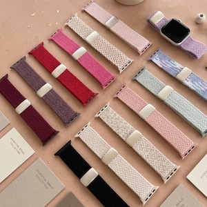 May include: A selection of braided watch straps in shades of pink, red, black, and purple. The straps are shown with a silver smartwatch and small cards. The straps are part of the "Essential Braid Collection."