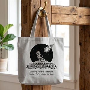 Puede incluir: Bolso tote gris claro con un gráfico negro de un extraterrestre mirando un cometa. El texto dice "RUSH RUSH RUSH" y "Waiting for the Asteroid. (Spoiler: You're missing the show.)". El bolso cuelga de un gancho dorado en una viga de madera.