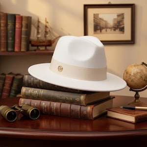 Könnte beinhalten: Ein weißer Fedora-Hut mit beigem Band und goldenem Emblem, der auf einem Stapel antiker Bücher liegt. Der Hut hat eine breite Krempe und eine klassische Form. Die Bücher sind ledergebunden mit goldener Schrift. Ein Fernglas ist ebenfalls zu sehen.