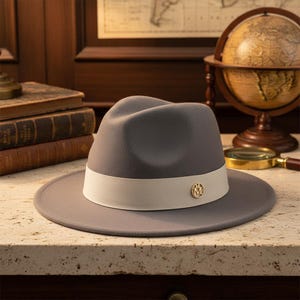 May include: A gray fedora hat with a cream-colored band and a gold-colored emblem. The hat is displayed on a light-colored surface. The hat's brim is approximately 3 inches wide.