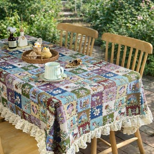 Puede incluir: Un mantel colorido con estampado de patchwork y ribete de encaje, colocado sobre una mesa de madera. El mantel presenta una variedad de cuadrados con motivos florales y de casas en tonos azules, verdes, morados y rojos. Una taza y un platillo están sobre la mesa.