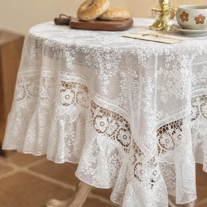 May include: A round, white lace tablecloth with a floral pattern and ruffled edge. The tablecloth is draped over a wooden table, with a wooden cutting board, bagels, and a teacup visible on top. The lace design adds a delicate, vintage aesthetic.