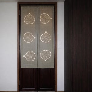 Puede incluir: Una cortina de puerta rectangular de tela beige con un estampado gr&aacute;fico blanco repetitivo de seis higos cortados por la mitad, dispuestos en dos columnas verticales. La cortina est&aacute; dividida en el centro y cuelga de un marco de puerta de madera oscura.